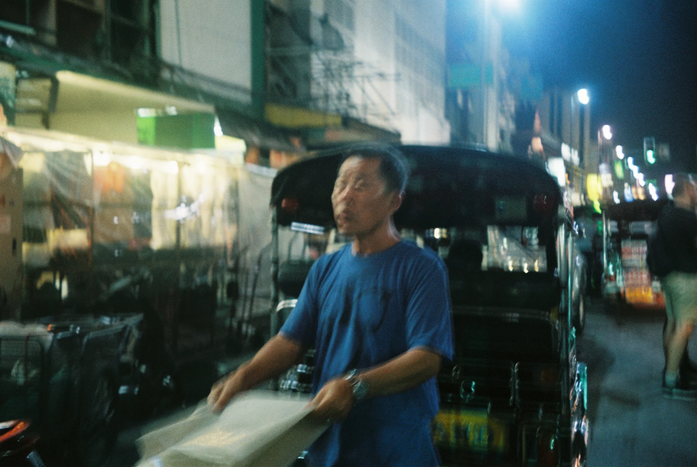 A man standing on a night street near tuk-tuks and market lights in Thailand.
